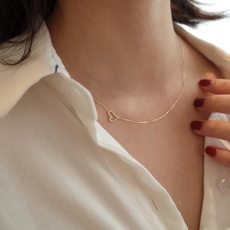 Close-up of a woman wearing a delicate gold necklace with a small open heart pendant, paired with a white blouse and dark red nail polish.