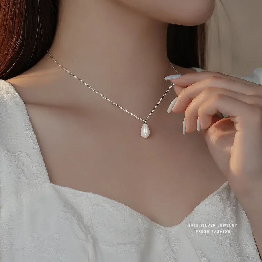 Close-up of a woman wearing a delicate silver necklace with a teardrop pearl pendant, paired with a textured white dress and long white manicured nails.