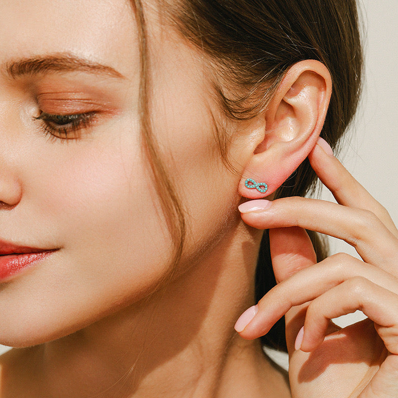 Close-up portrait of a woman wearing turquoise infinity-shaped stud earrings while gently touching her ear with manicured fingers.