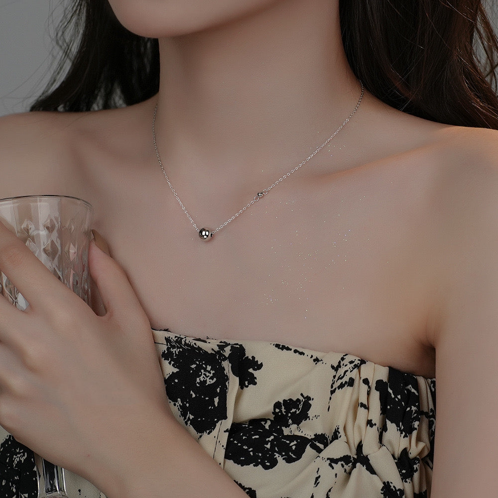 Close-up of a woman wearing a minimalist silver necklace with a small bead pendant while holding a textured glass and wearing a strapless patterned top.