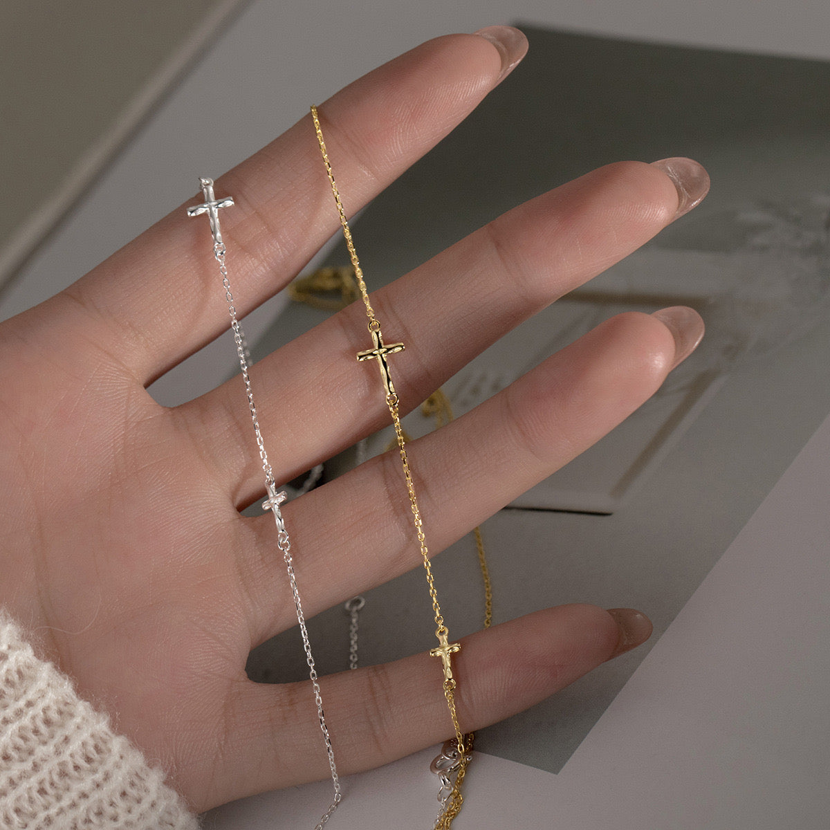 Close-up of a hand holding delicate gold and silver chain bracelets featuring small cross charms against a soft neutral background.