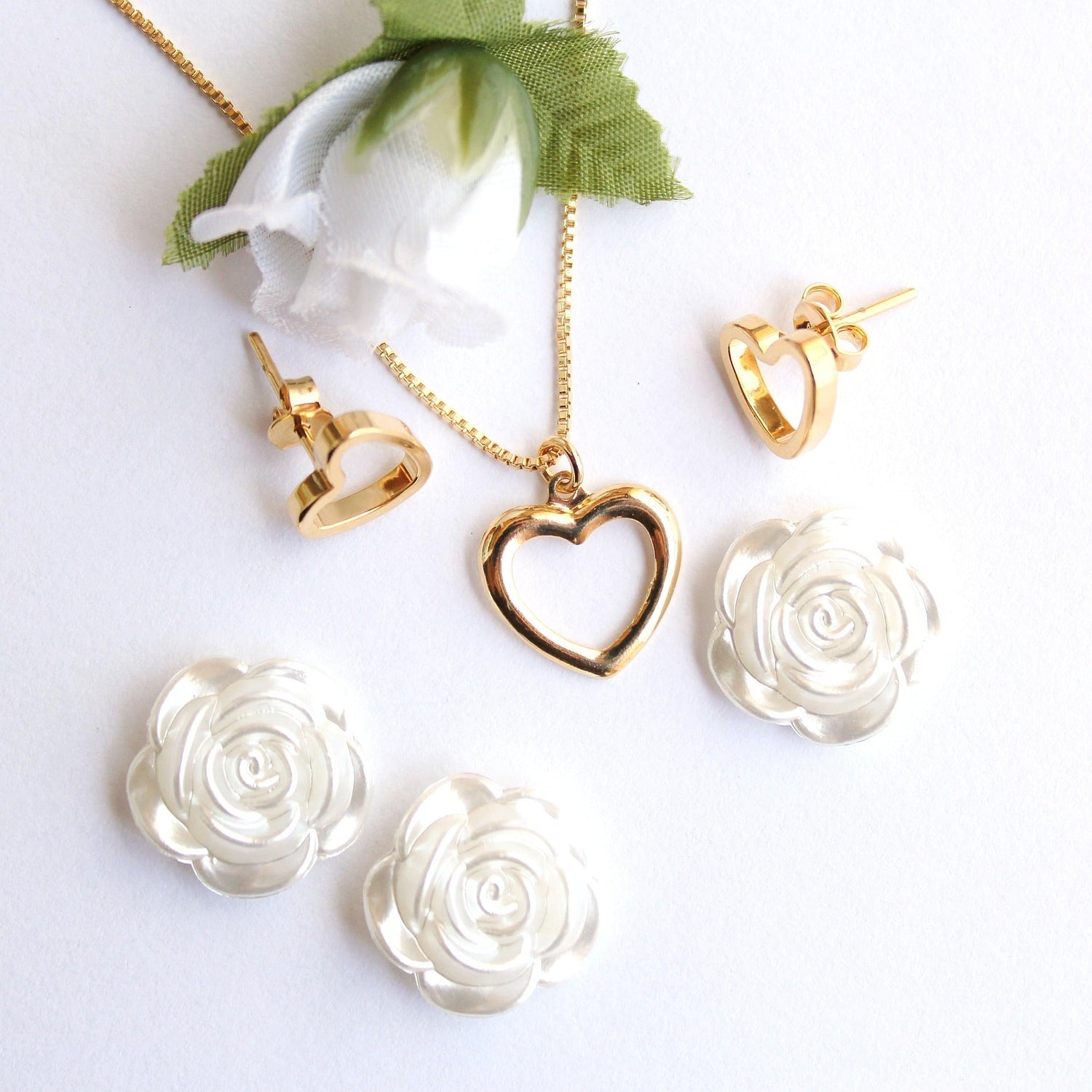 Gold heart-shaped necklace and matching earrings displayed with white floral accents on a bright white background.