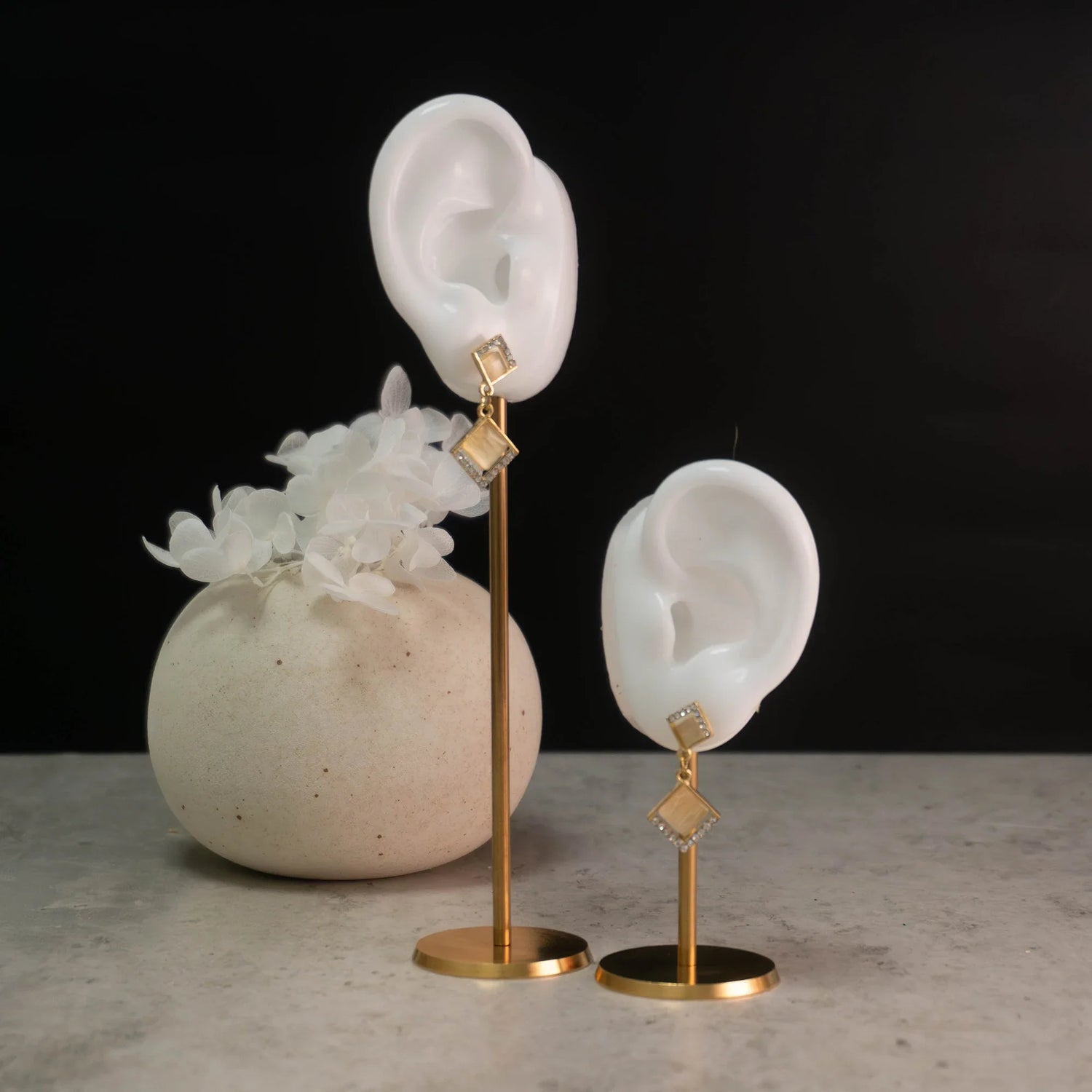 Gold geometric drop earrings displayed on white ear stands beside a vase of white flowers.