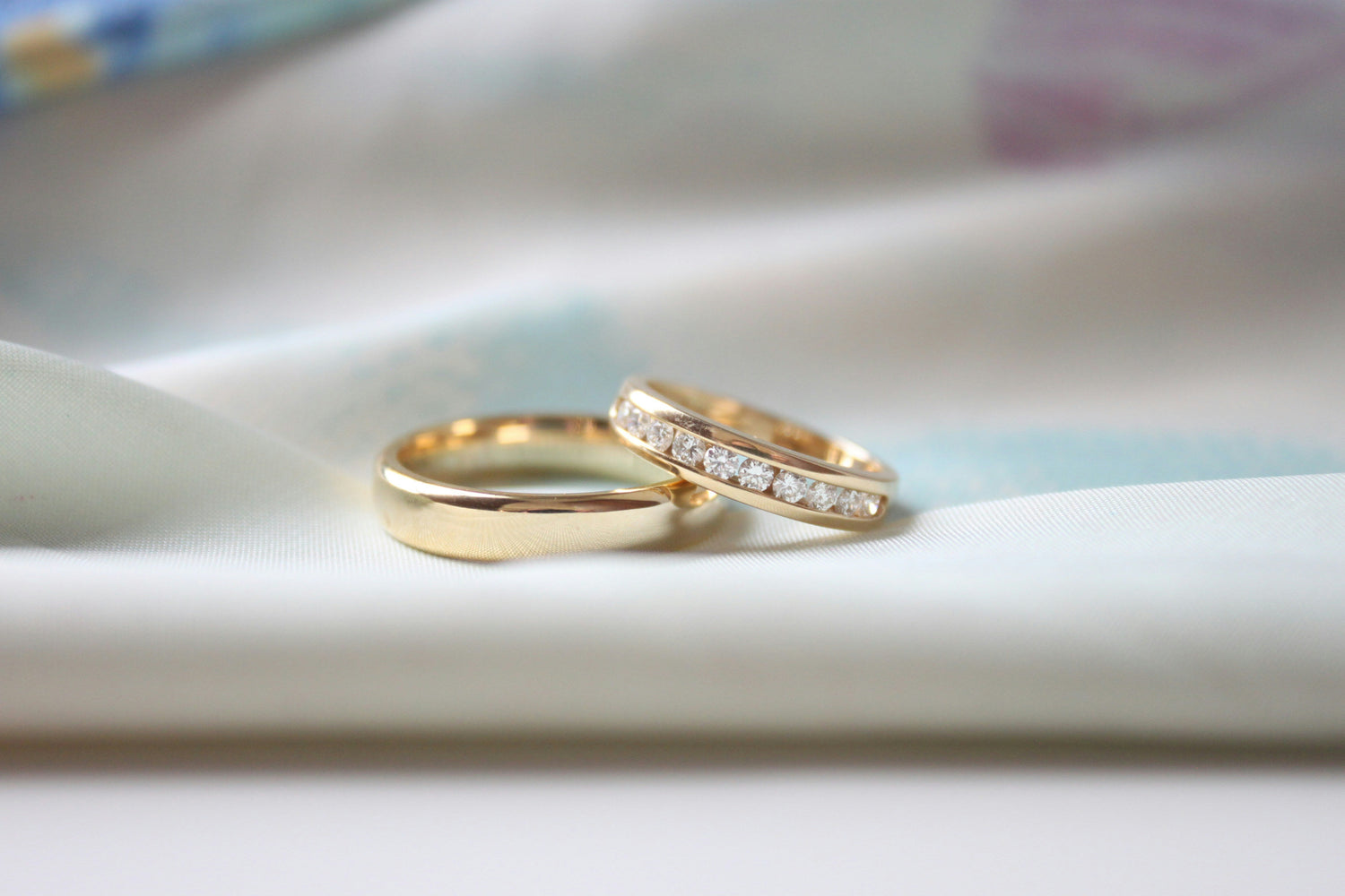 Two gold rings resting on soft white fabric, including a plain gold band and a diamond-studded eternity ring in close-up focus.