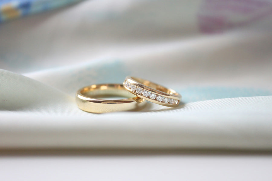 Two gold rings resting on soft white fabric, including a plain gold band and a diamond-studded eternity ring in close-up focus.