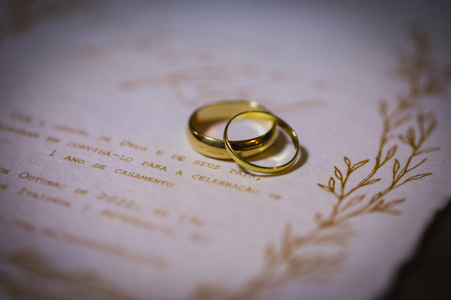 Close-up of two gold wedding bands resting on an elegant wedding invitation with soft lighting, shallow depth of field, and romantic decorative details in the background.