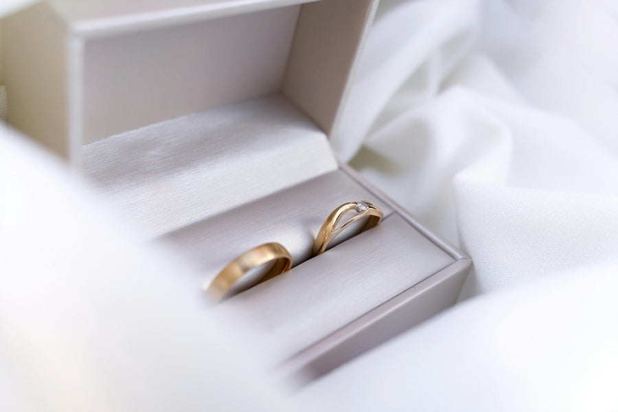 Close-up of two gold wedding rings inside a white jewelry box, surrounded by soft white fabric with a clean, elegant, and romantic aesthetic.