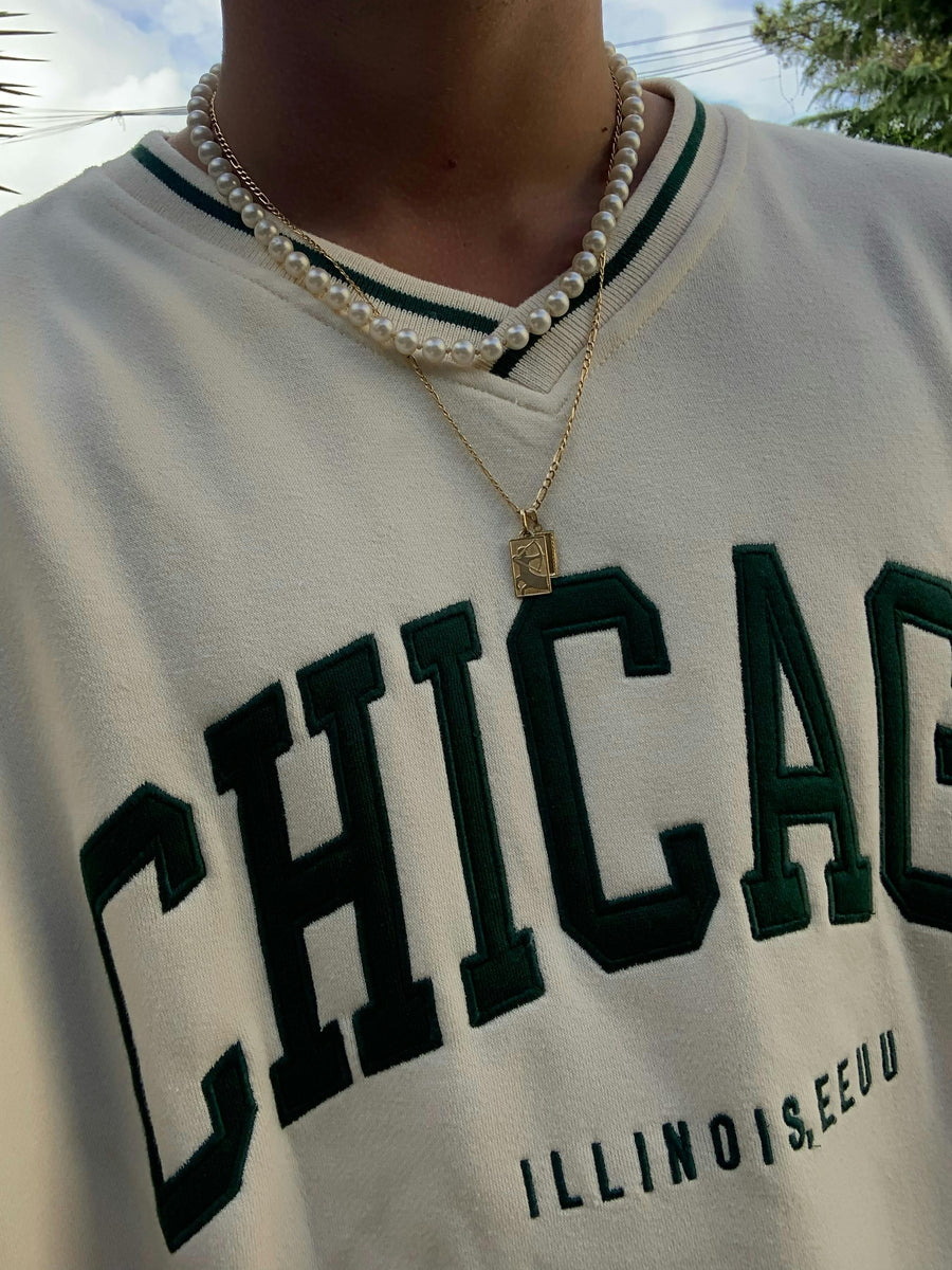 Close-up of layered pearl and gold necklaces worn with a white Chicago sweatshirt outdoors in natural light.
