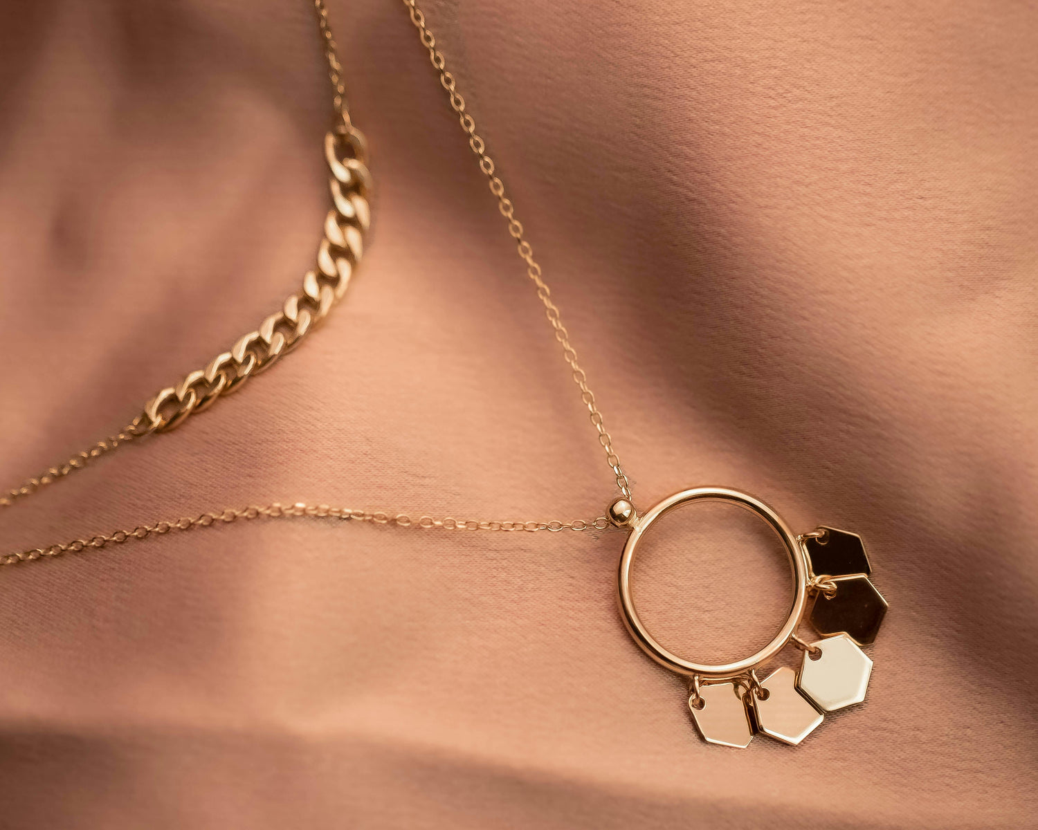 Close-up of layered gold necklaces with a circular pendant and geometric charms against warm-toned fabric.