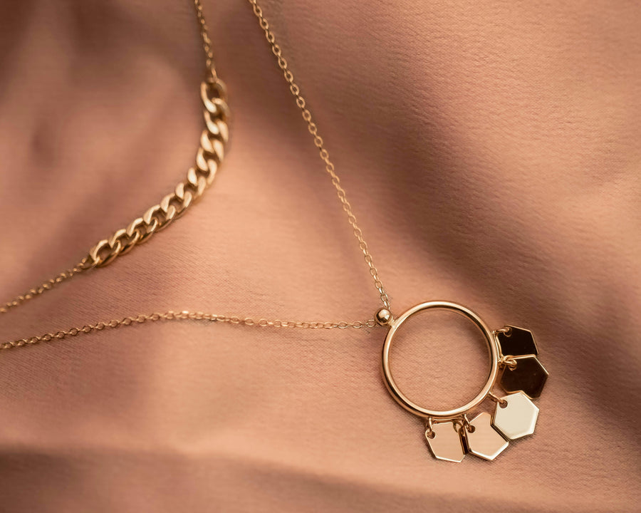 Close-up of layered gold necklaces with a circular pendant and geometric charms against warm-toned fabric.