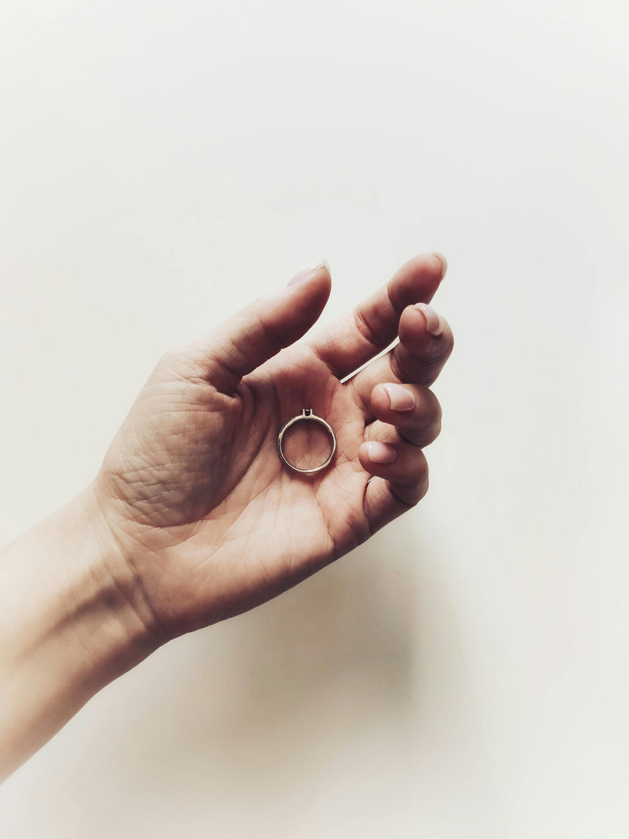 Close-up of a hand holding a simple ring against a soft white background with minimal lighting and a delicate, emotional aesthetic.