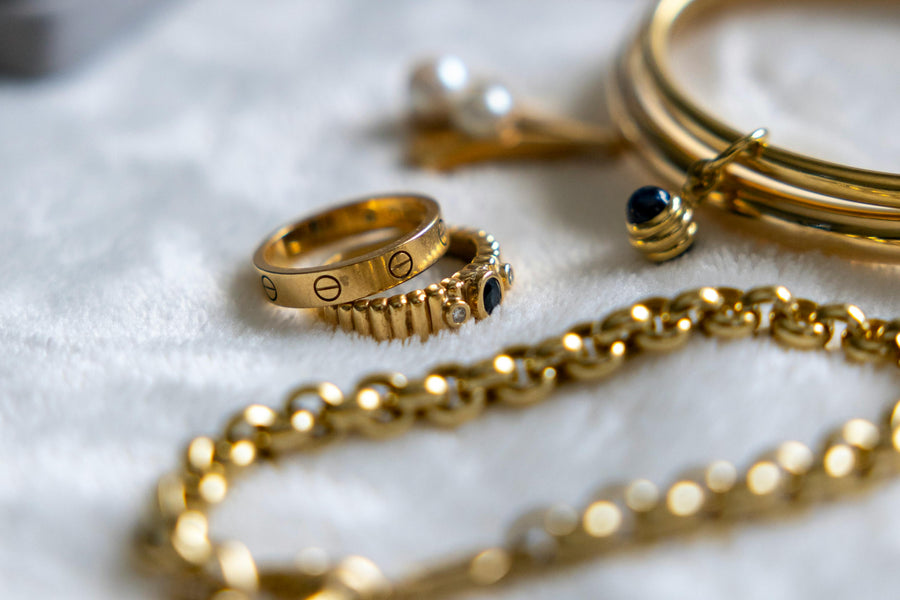 Close-up of gold jewelry including stacked rings, bracelets, and a chain resting on a soft white fabric surface with a blurred background.