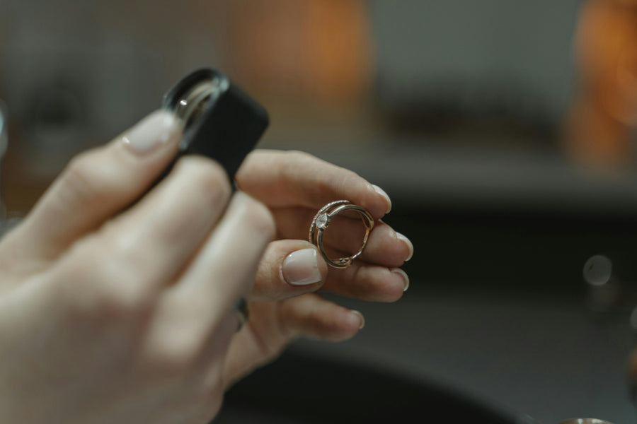 Close-up of hands examining a diamond ring with a jewelry loupe in a softly lit setting, featuring a blurred background and elegant, intimate atmosphere.